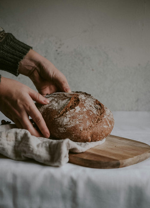 Person holding a loaf of bread on a wooden cutting board with a textured wall background