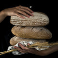 Hands holding three loaves of bread and a bundle of wheat against a black background