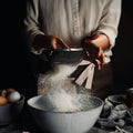 Person sifting flour into a bowl with eggs and other ingredients on a dark surface