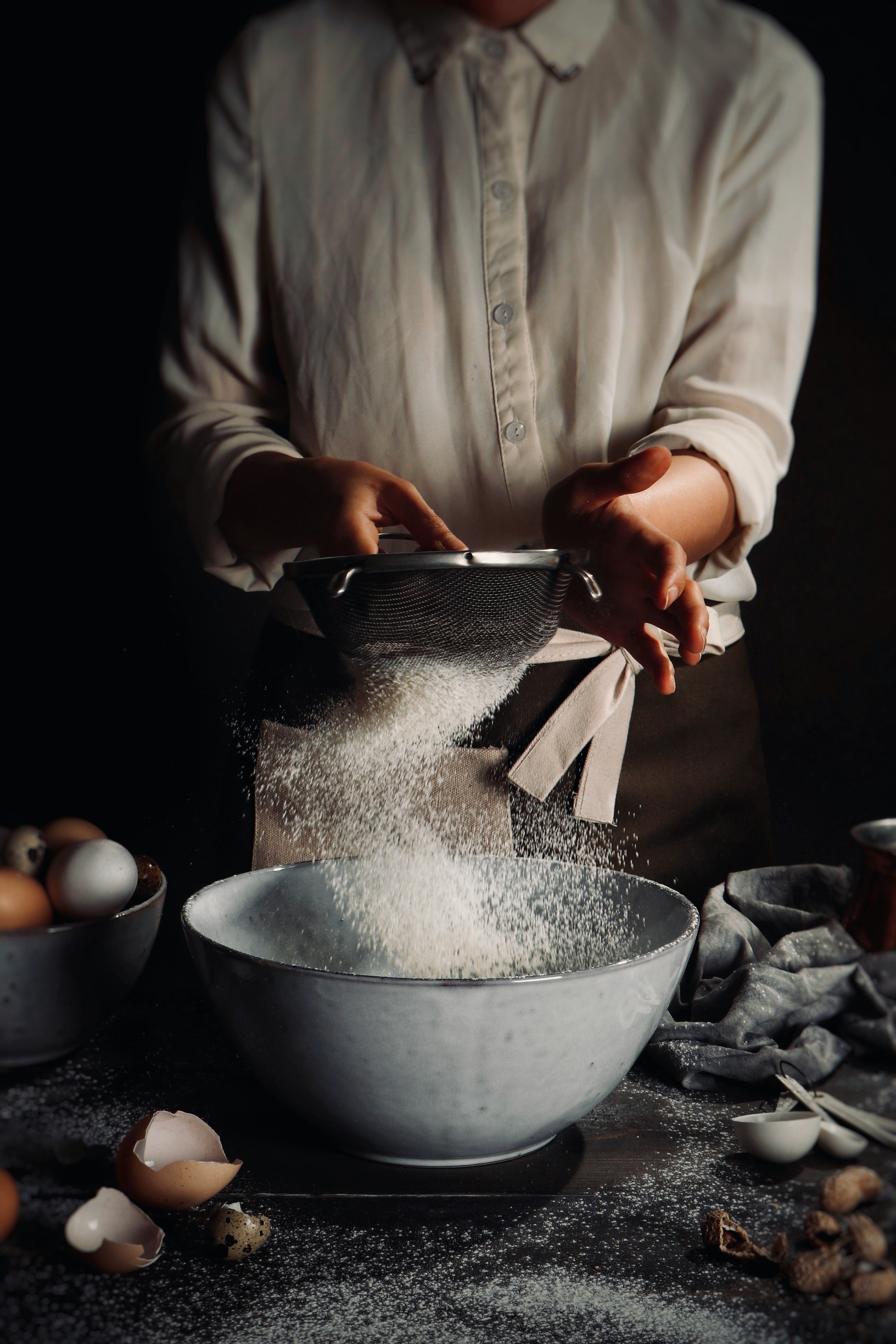 Person sifting flour into a bowl with eggs and other ingredients on a dark surface