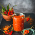 Glass jar of red chili paste with chili peppers and a spoonful on a dark surface
