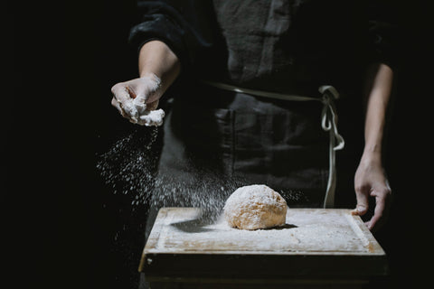 A baker wearing a dark apron sprinkles flour over a round piece of dough resting on a wooden board against a dark background.
