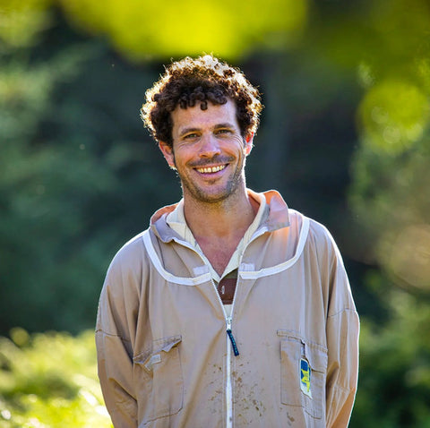 A smiling man outdoors wearing a beige zip-front hooded jacket with white trim over a white shirt, set against a blurred green natural background.